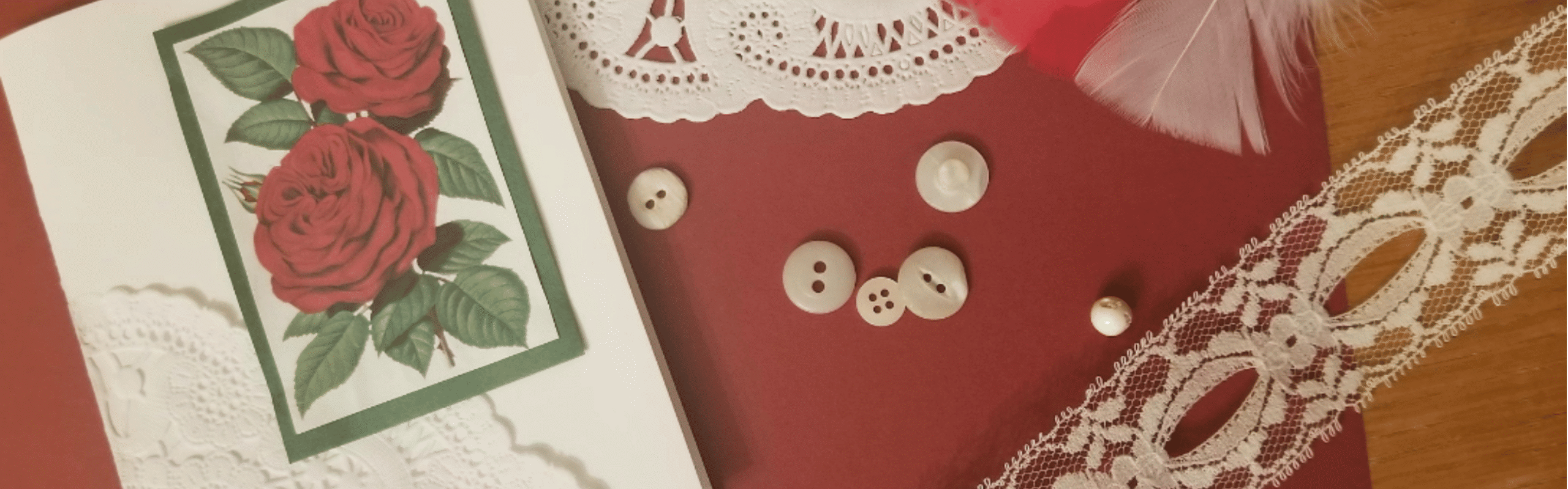 Craft supplies including a rose card, white buttons, lace, and a feather on red and wood surfaces.